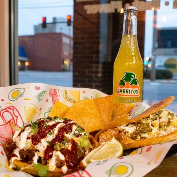Mexican street food basket with a Jarritos pineapple soda, tacos, and nachos by a window.