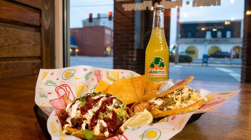 Mexican street food basket with a Jarritos pineapple soda, tacos, and nachos by a window.