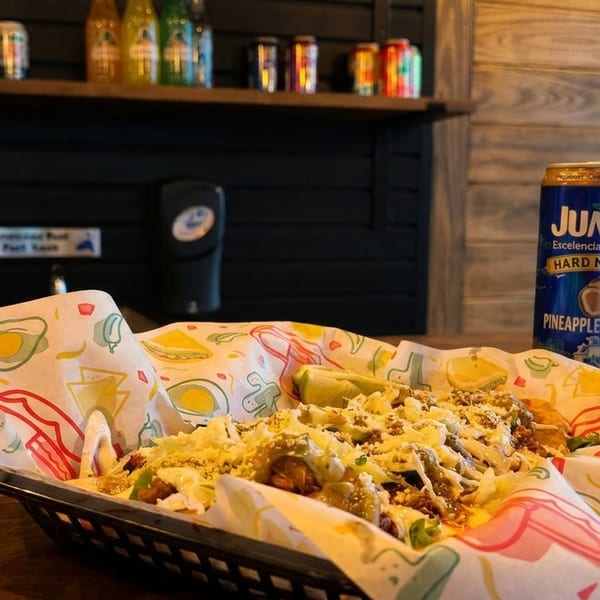 A basket of tacos with shredded lettuce, cheese, and lime, next to a Jumex pineapple coconut drink.