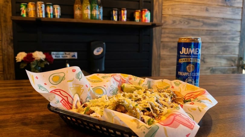 A basket of tacos with shredded lettuce, cheese, and lime, next to a Jumex pineapple coconut drink.