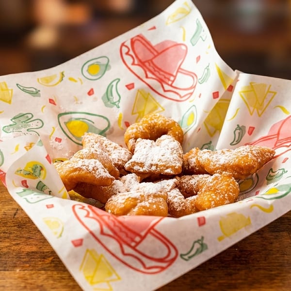 Golden brown churros dusted with powdered sugar in a basket with festive Mexican-themed paper.
