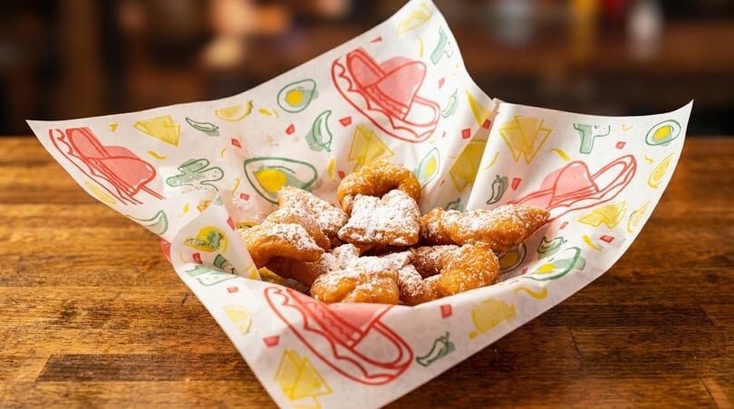 Golden brown churros dusted with powdered sugar in a basket with festive Mexican-themed paper.