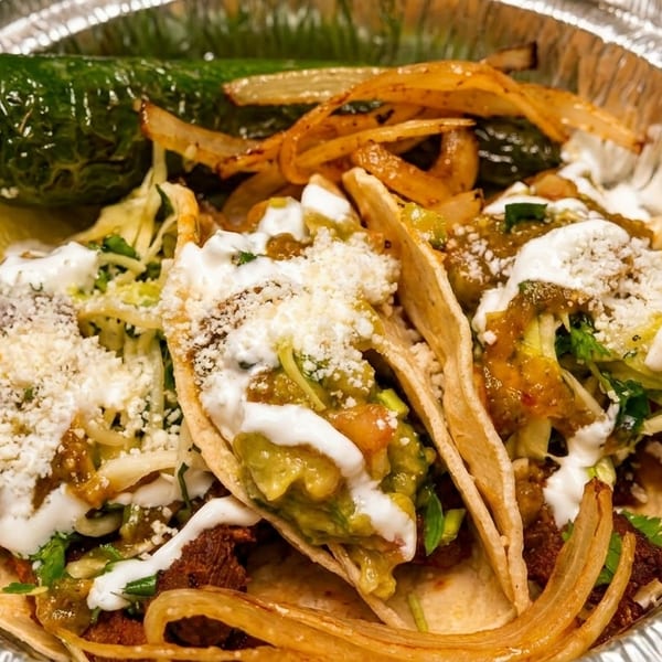 Three tacos with guacamole, sour cream, cheese, and grilled onions in a foil container.