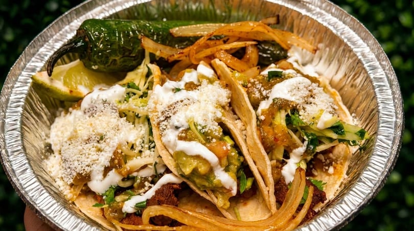 Three tacos with guacamole, sour cream, cheese, and grilled onions in a foil container.