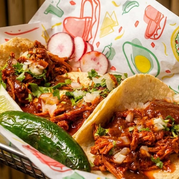 Two birria tacos with cilantro, onions, lime, jalapeño, and radishes in a basket.