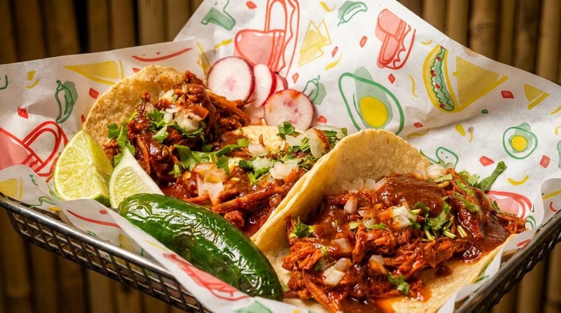 Two birria tacos with cilantro, onions, lime, jalapeño, and radishes in a basket.