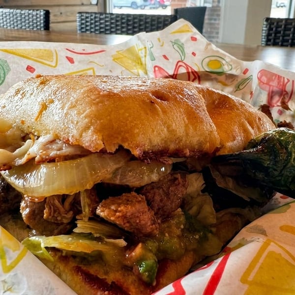 A close-up of a hearty torta sandwich with meat, onions, and guacamole, served in colorful paper.