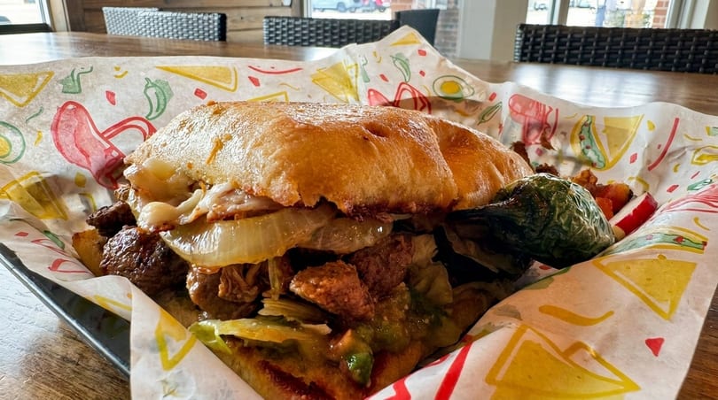 A close-up of a hearty torta sandwich with meat, onions, and guacamole, served in colorful paper.